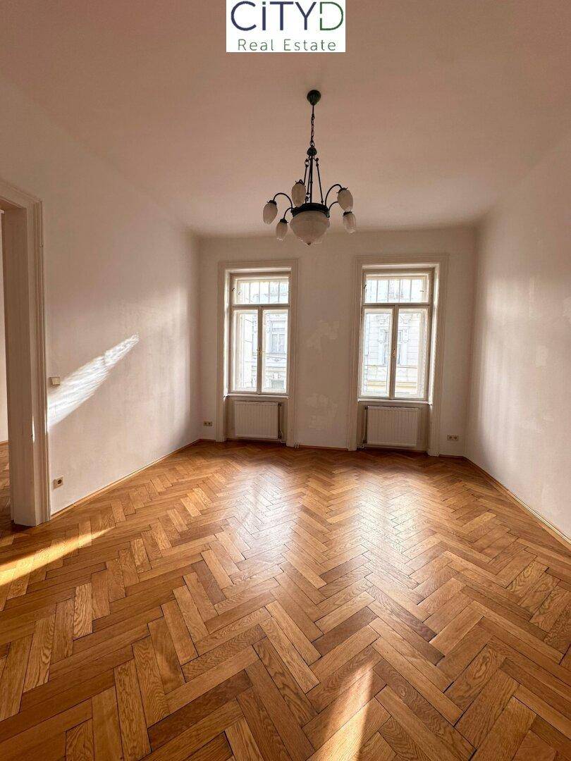 Geräumiges Zimmer mit klassischem Fischgrätparkett, Kronleuchter und zwei hohen Fenstern.