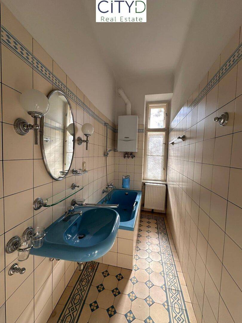 Badezimmer im 80er-Jahre-Stil mit blauer Badewanne, Waschbecken und gemusterten Fliesen.