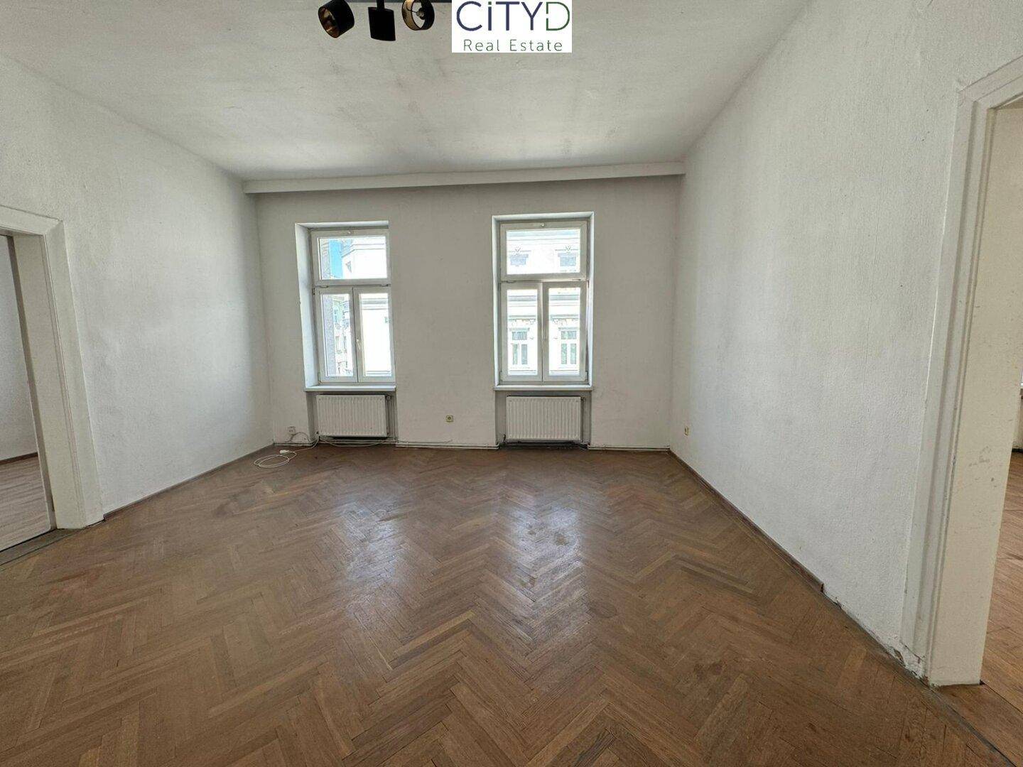 Helles Zimmer mit Fischgrätparkett und zwei Fenstern, bereit zur Renovierung.