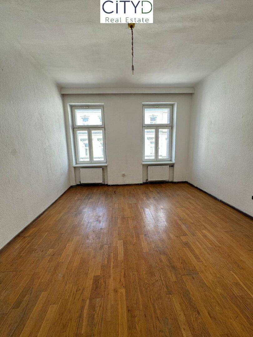 Leeres Zimmer mit Holzboden und zwei Fenstern, wartet auf neue Bewohner.