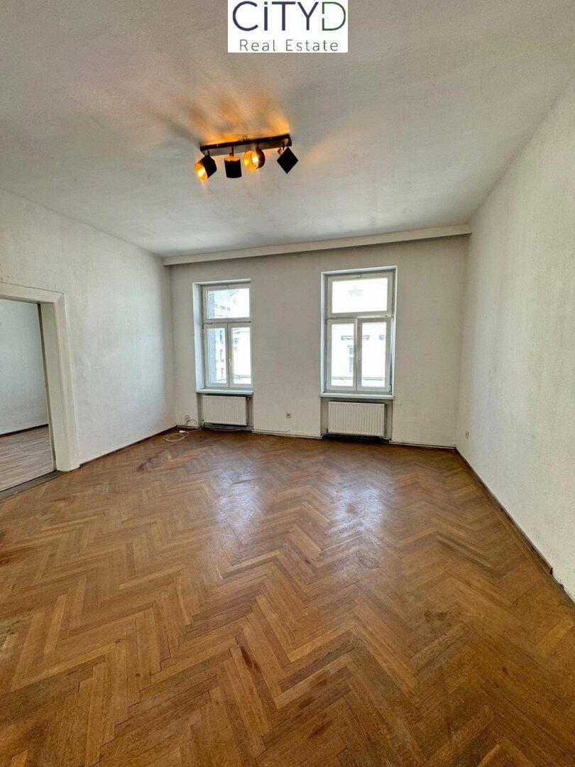 Geräumiges Zimmer mit Fischgrätparkett und zwei Fenstern, ideal für Wohnzwecke.