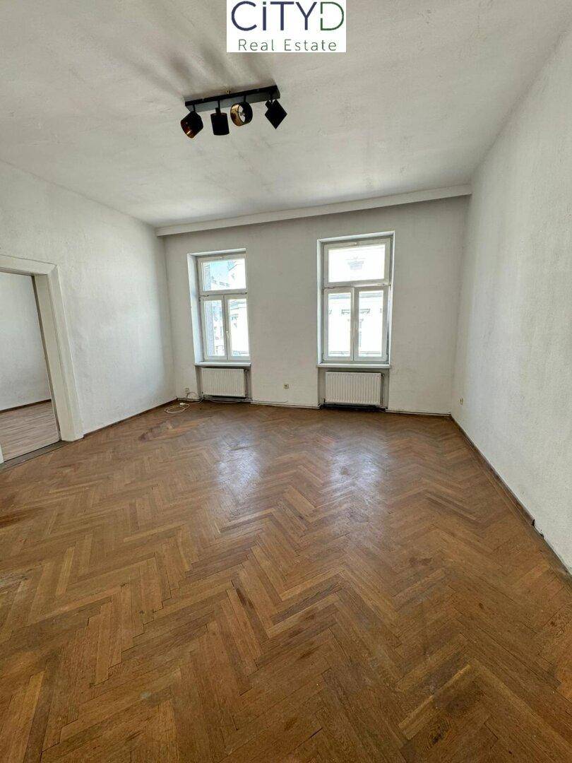 Helles Zimmer mit Fischgrätparkett und zwei Fenstern, bereit zur Renovierung.