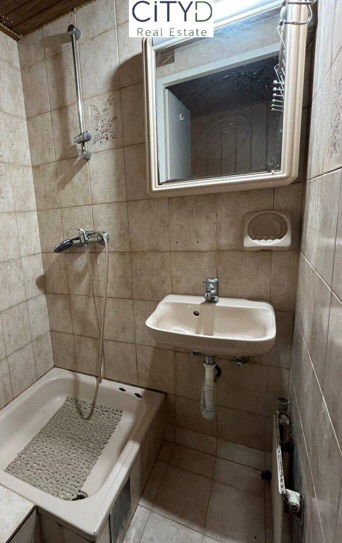 Kleines Badezimmer mit Badewanne, Dusche und Waschbecken, gefliest und renovierungsbedürftig.