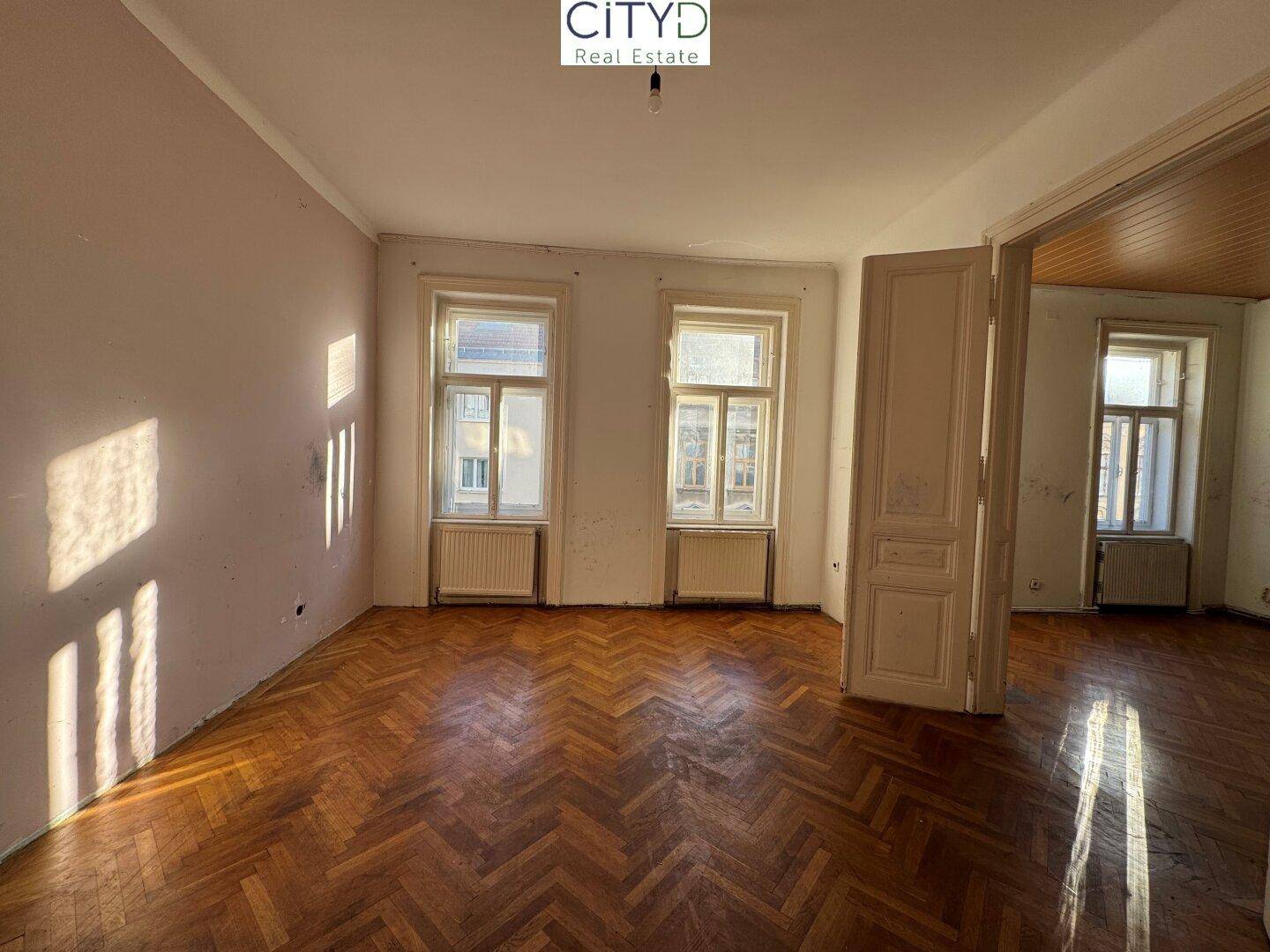 Helles Wohnzimmer mit Fischgrätparkett und Blick in den Nebenraum, renovierungsbedürftig.