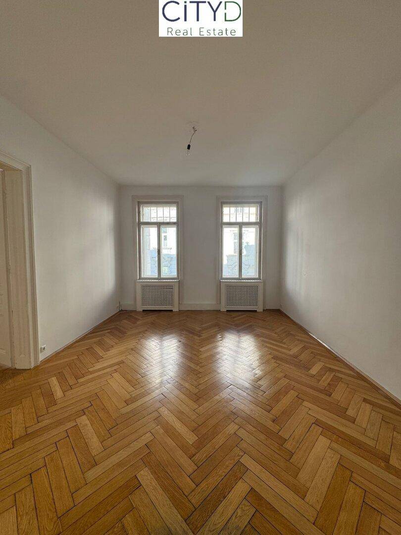 Heller, leerer Raum mit klassischem Fischgrätparkett und zwei großen Fenstern.
