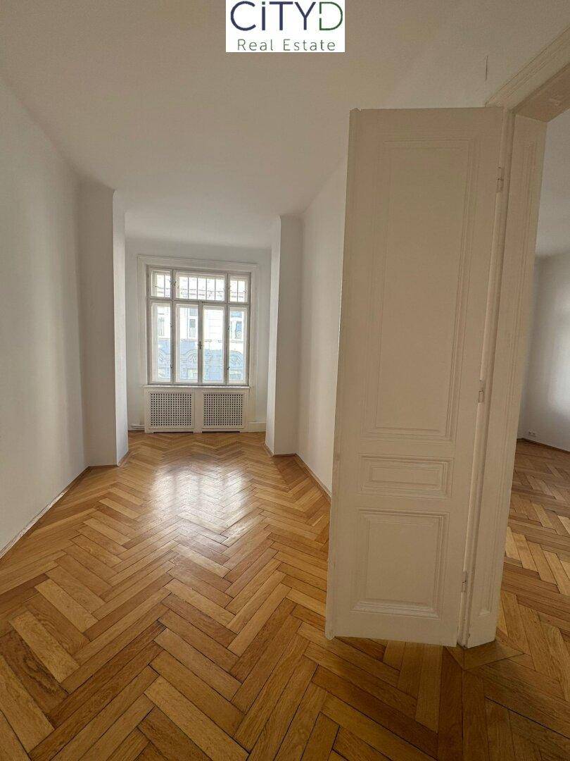 Geräumiger, leerer Raum mit Fischgrätparkett und einer hohen Flügeltür.