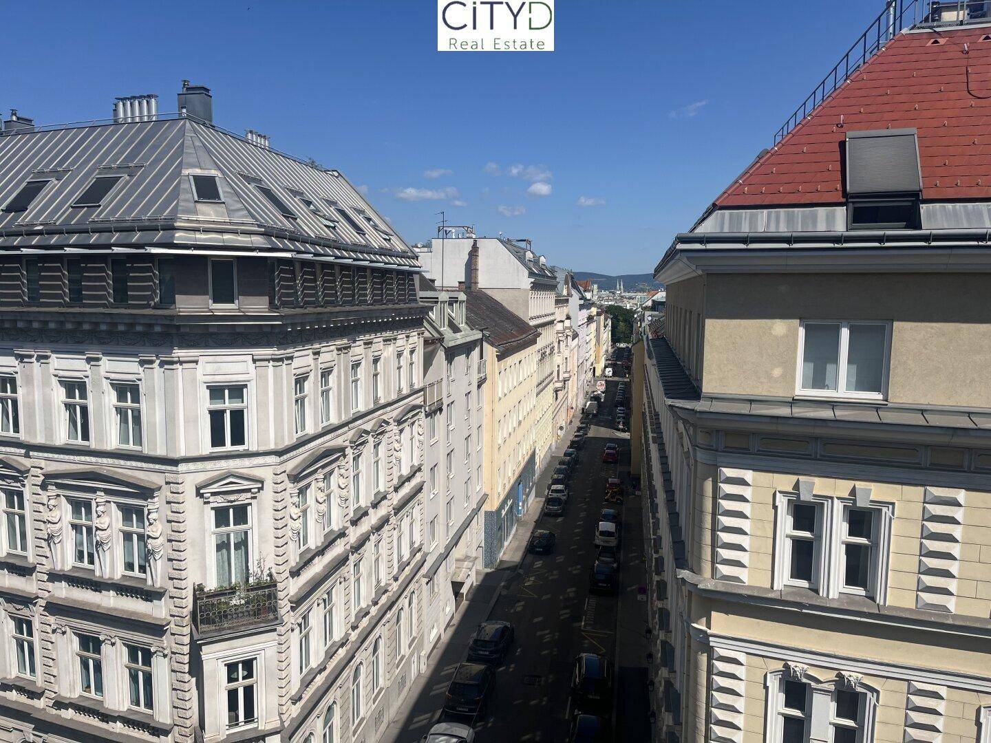Blick von oben auf eine belebte Stadtstraße mit historischen Gebäuden und parkenden Autos unter blauem Himmel.
