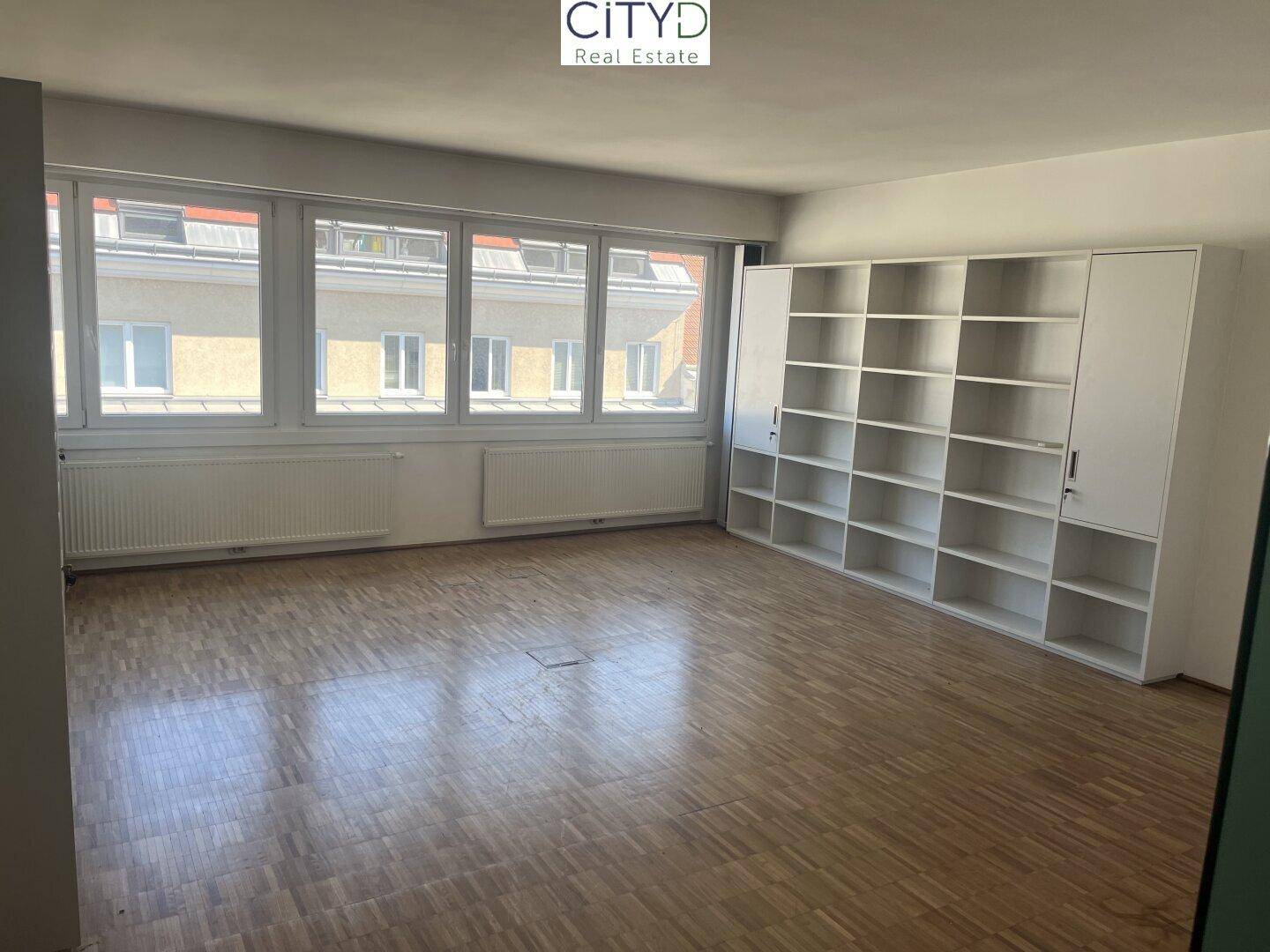 Geräumiger Büroraum mit großer Regalwand, hellen Fenstern und klassischem Parkettboden, bietet viel Stauraum.