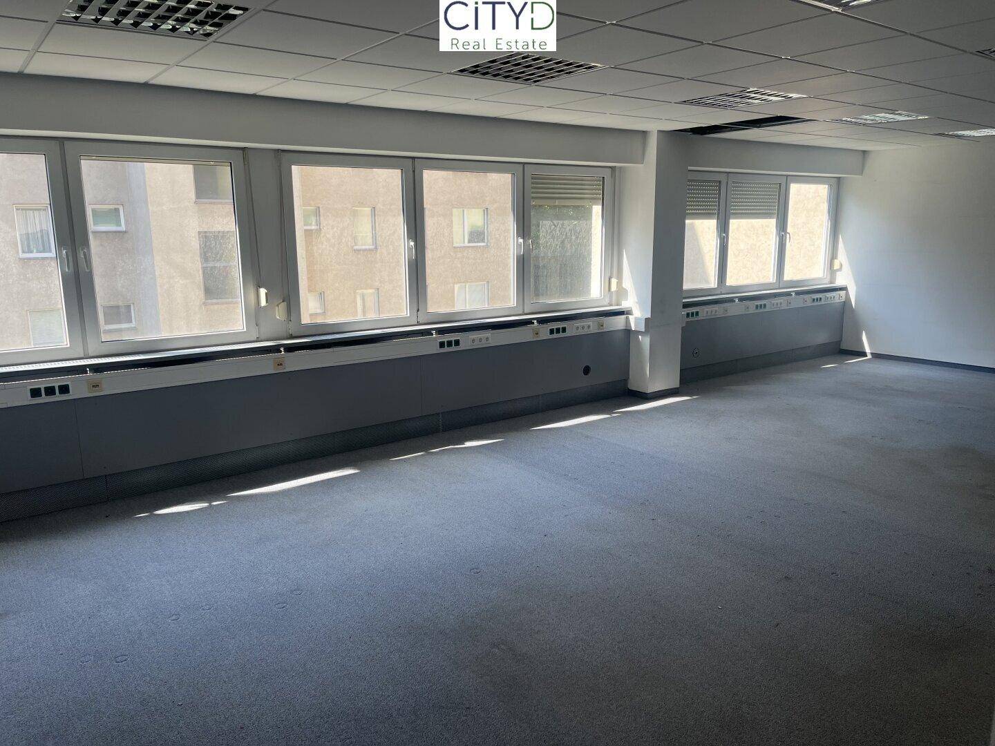 Geräumiges, leeres Büro mit Teppichboden, großen Fenstern und Deckenbeleuchtung.