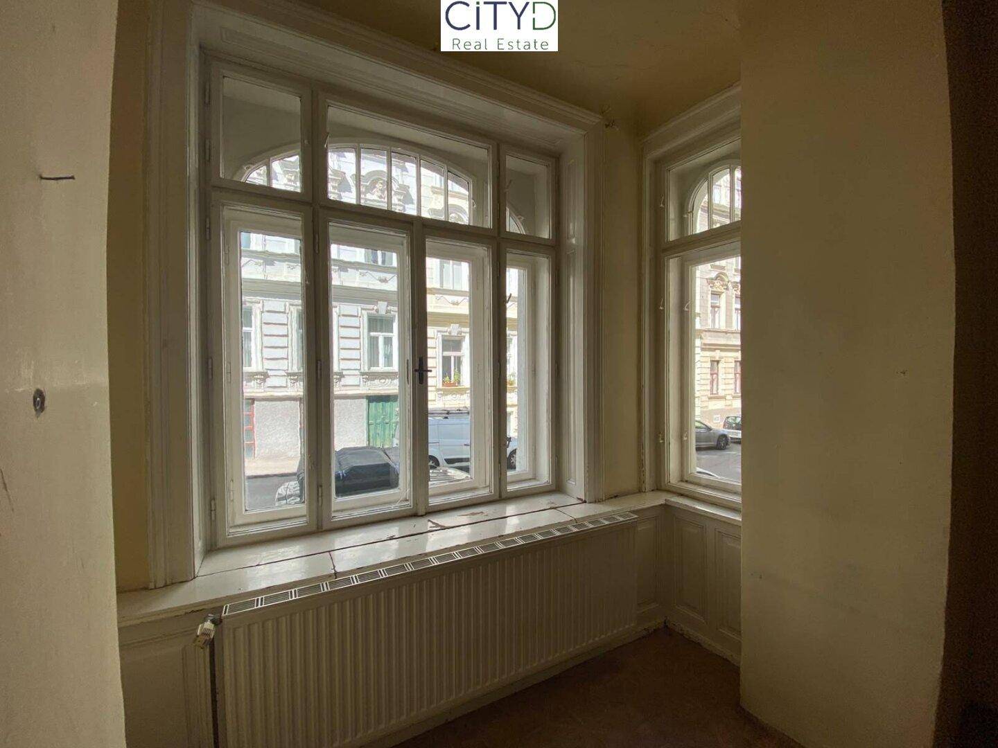 Helles Zimmer mit großen, historischen Fenstern, Heizkörper und Blick auf die Straße.