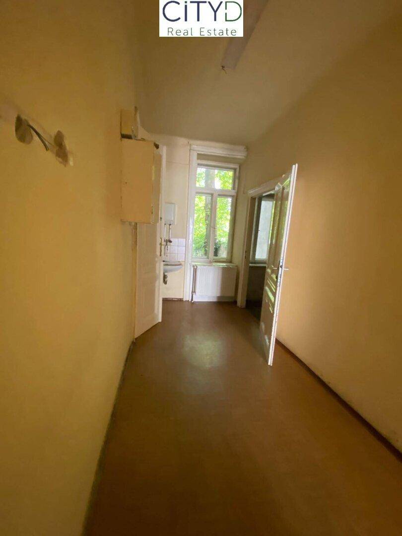 Langer Flur mit gelben Wänden, braunem Boden und Zugang zu einem Raum mit Fenster und Spülbecken.