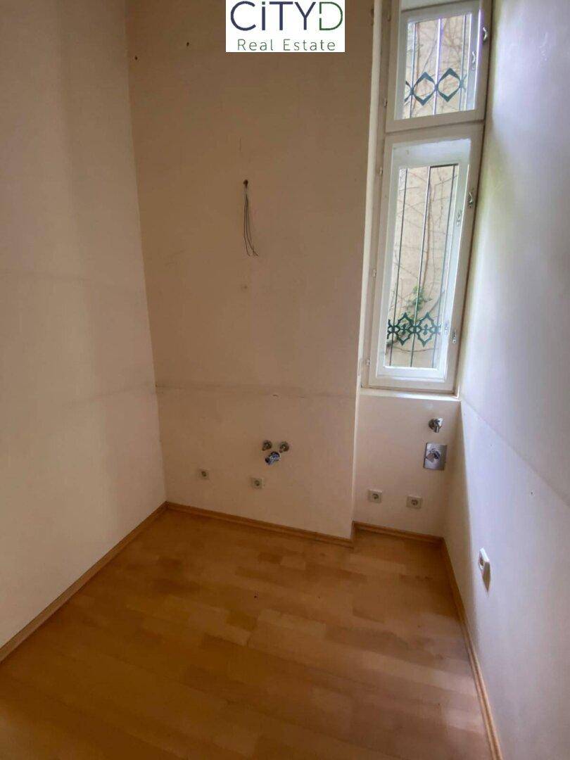 Kleiner Raum mit hellen Wänden, Holzfußboden und Fenster, vorbereitet für eine Küche.