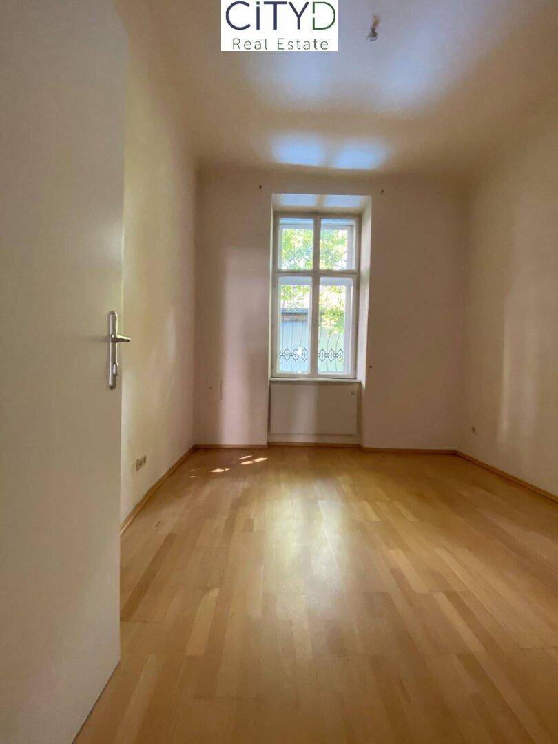 Helles, leeres Zimmer mit Holzfußboden und großem Fenster, ideal als Schlaf- oder Wohnbereich.