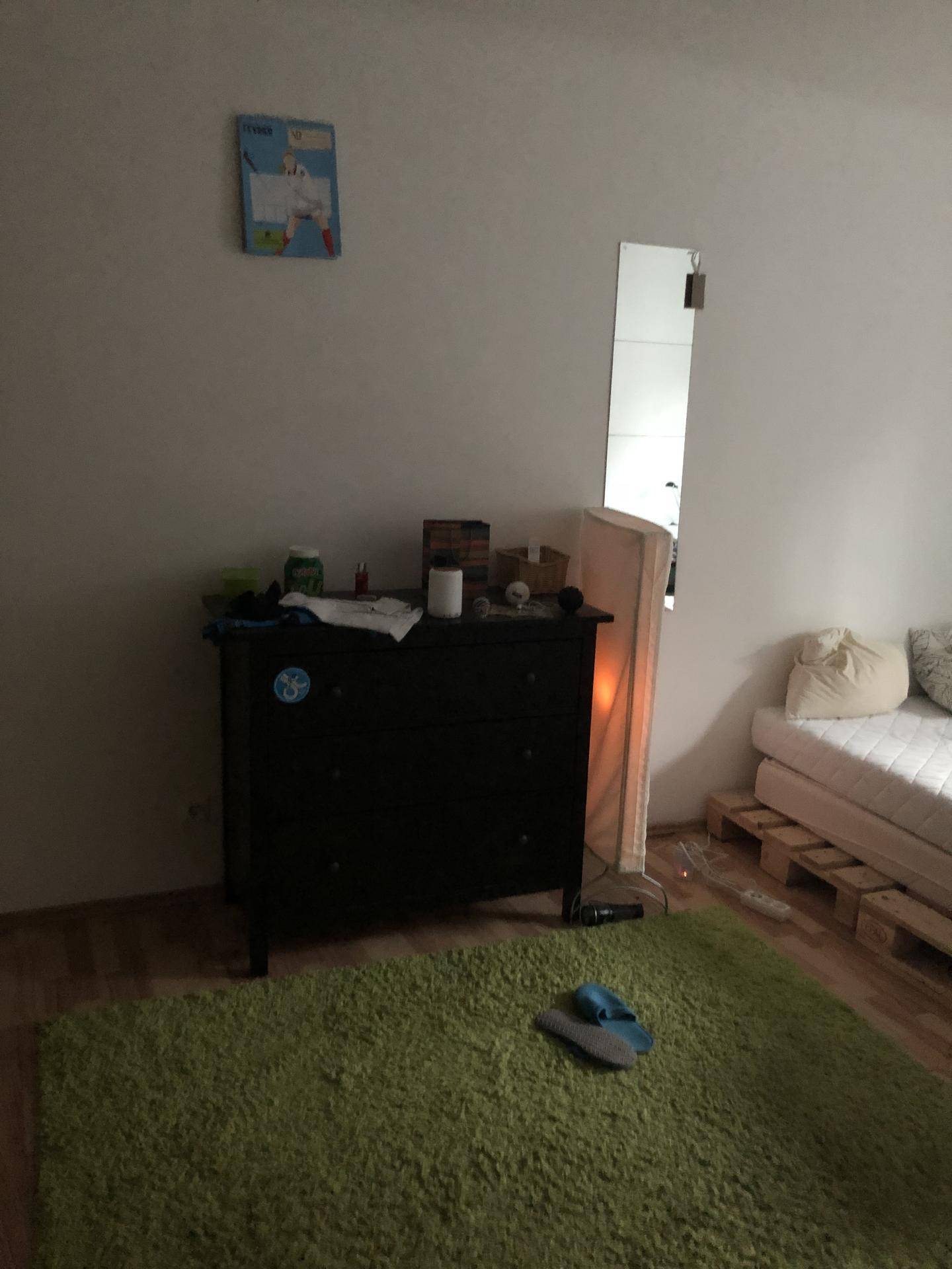 Schlafzimmer mit dunkler Kommode und Bett auf Palettenbasis, beleuchtet durch eine Stehlampe.