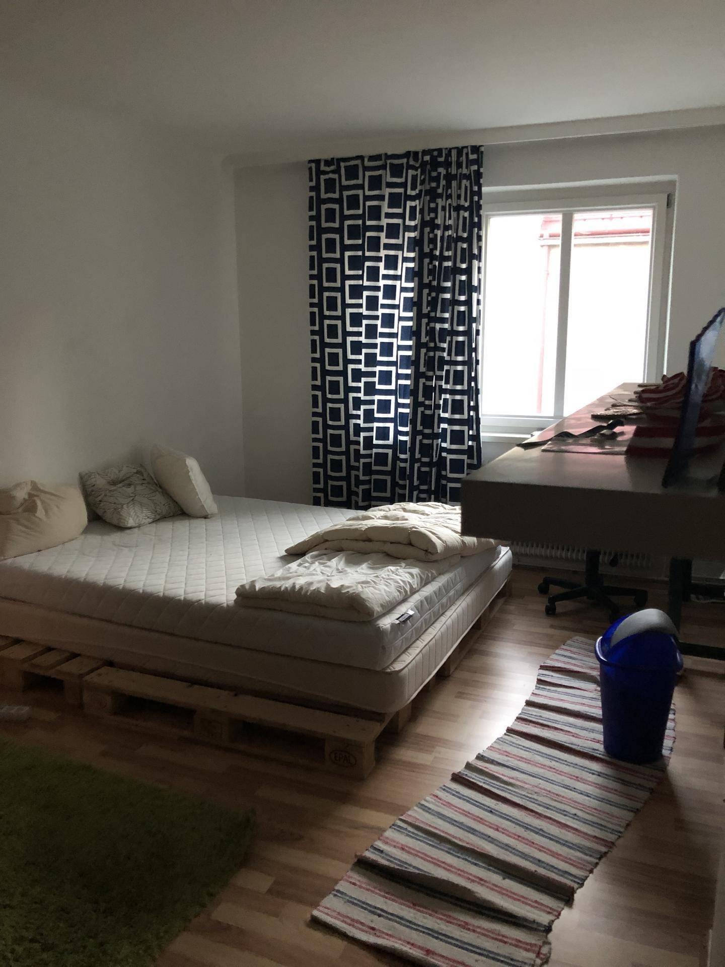 Schlafzimmer mit Bett auf Palettenbasis, Holzboden und Fenster mit gemusterten Vorhängen.