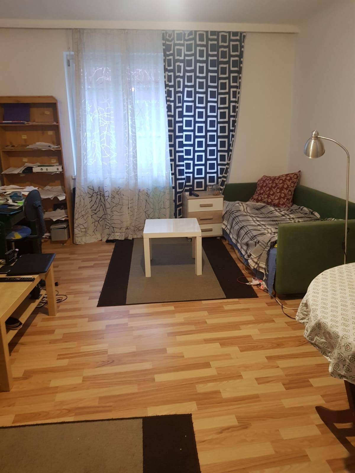 Wohnzimmer mit Tagesbett, Bücherregal und Couchtisch, beleuchtet durch ein Fenster.