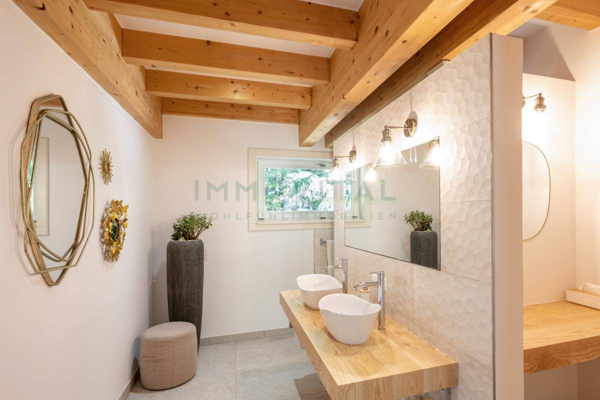 Modernes Badezimmer mit freiliegenden Holzbalken, Doppelwaschbecken auf Holzplatte und strukturierten Fliesen.