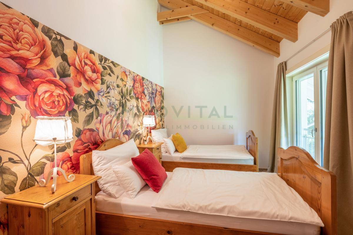 Helles Schlafzimmer mit floraler Tapete, Holzbalkendecke und zwei Einzelbetten.