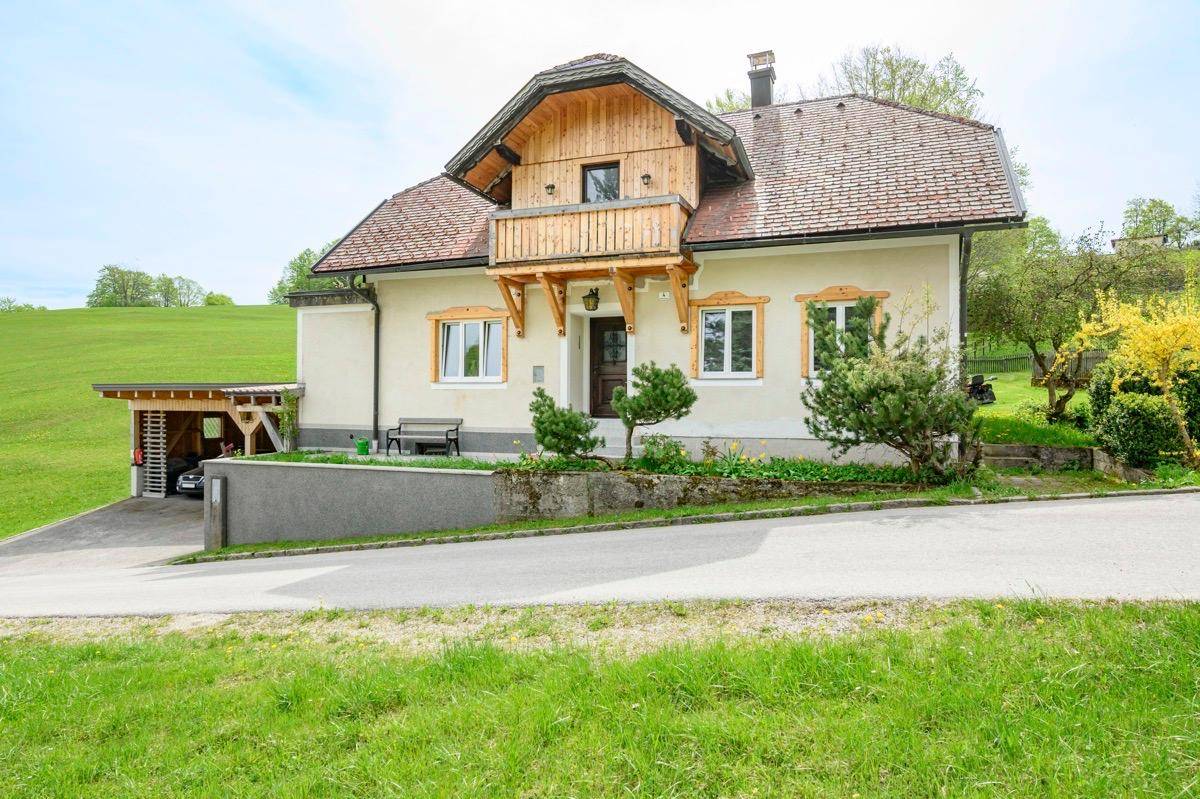 Charmantes Landhaus mit Holzbalkon und gepflegtem Vorgarten, umgeben von grünen Hügeln.