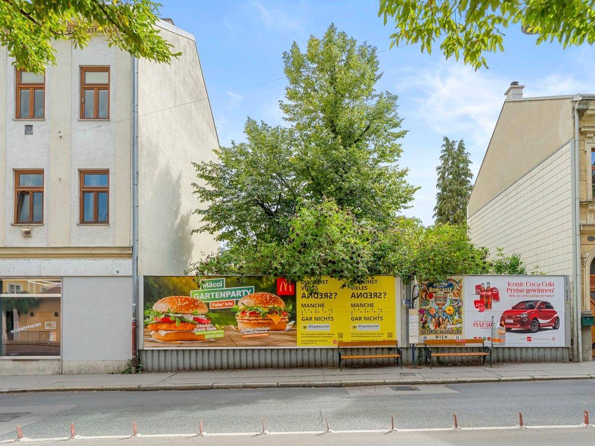 Straßenansicht mit Gebäuden, einem großen Baum und Werbetafeln, die die Umgebung prägen.