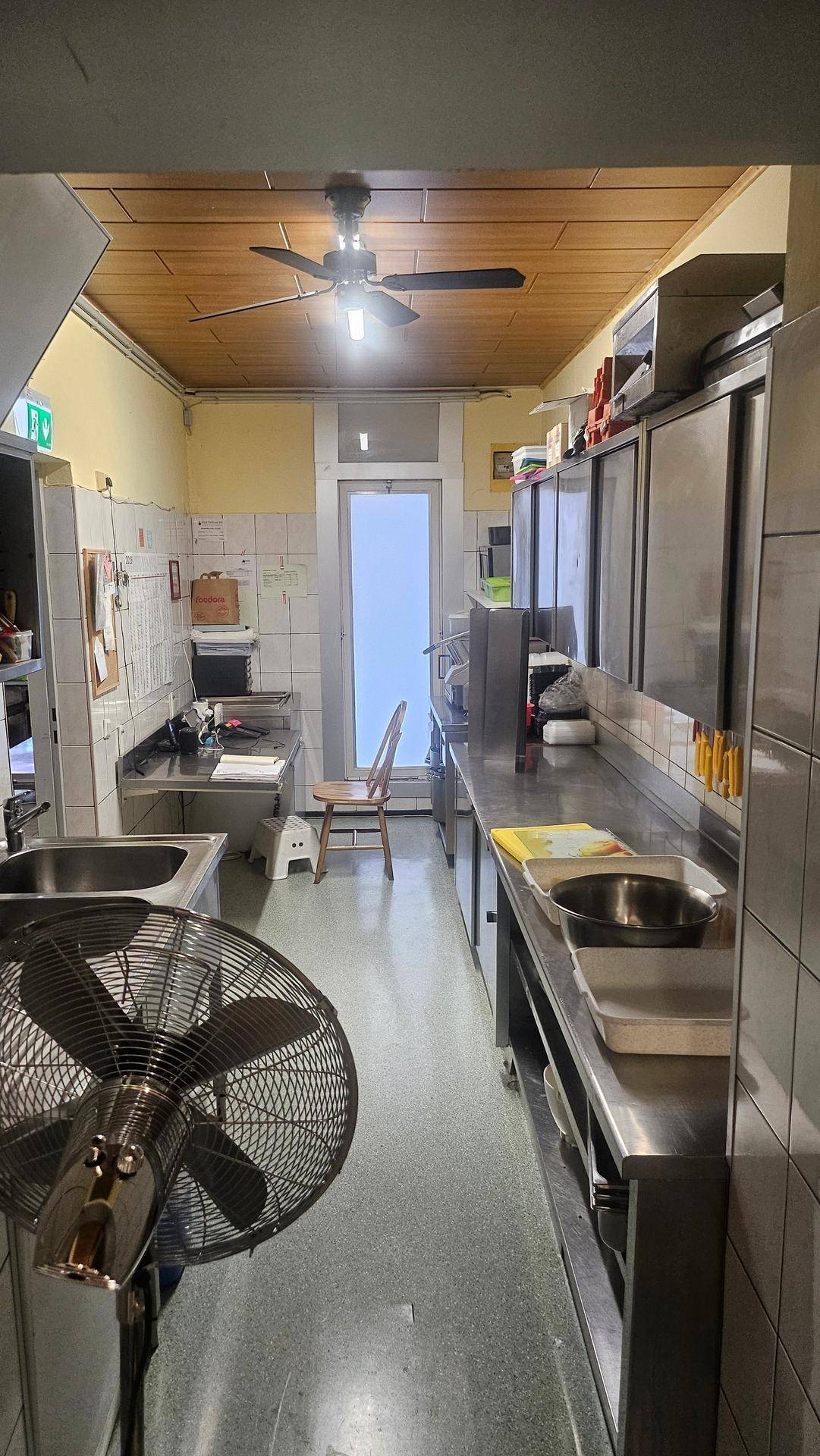Schmale Gewerbeküche mit Edelstahl-Arbeitsflächen, Deckenventilator und funktionaler Ausstattung.