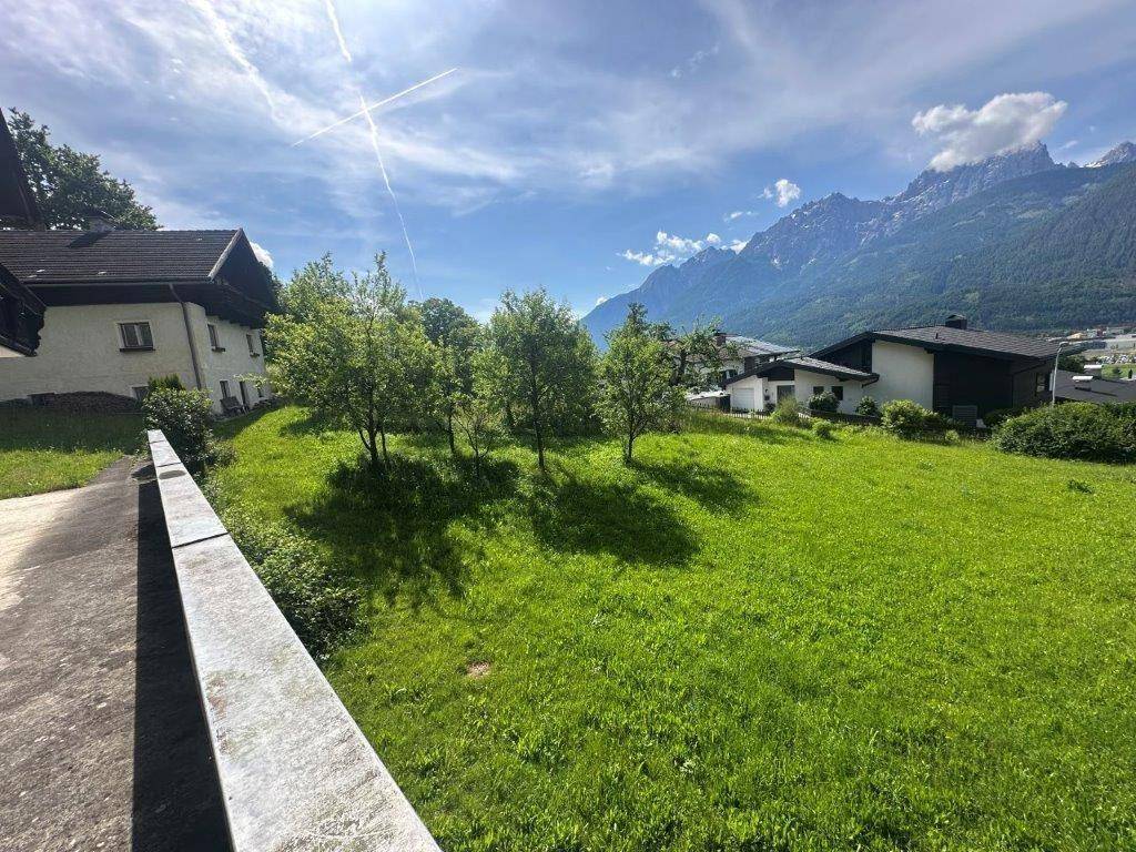 Weitläufige grüne Wiese mit vereinzelten Bäumen und Häusern, umrahmt von einer beeindruckenden Bergkulisse.
