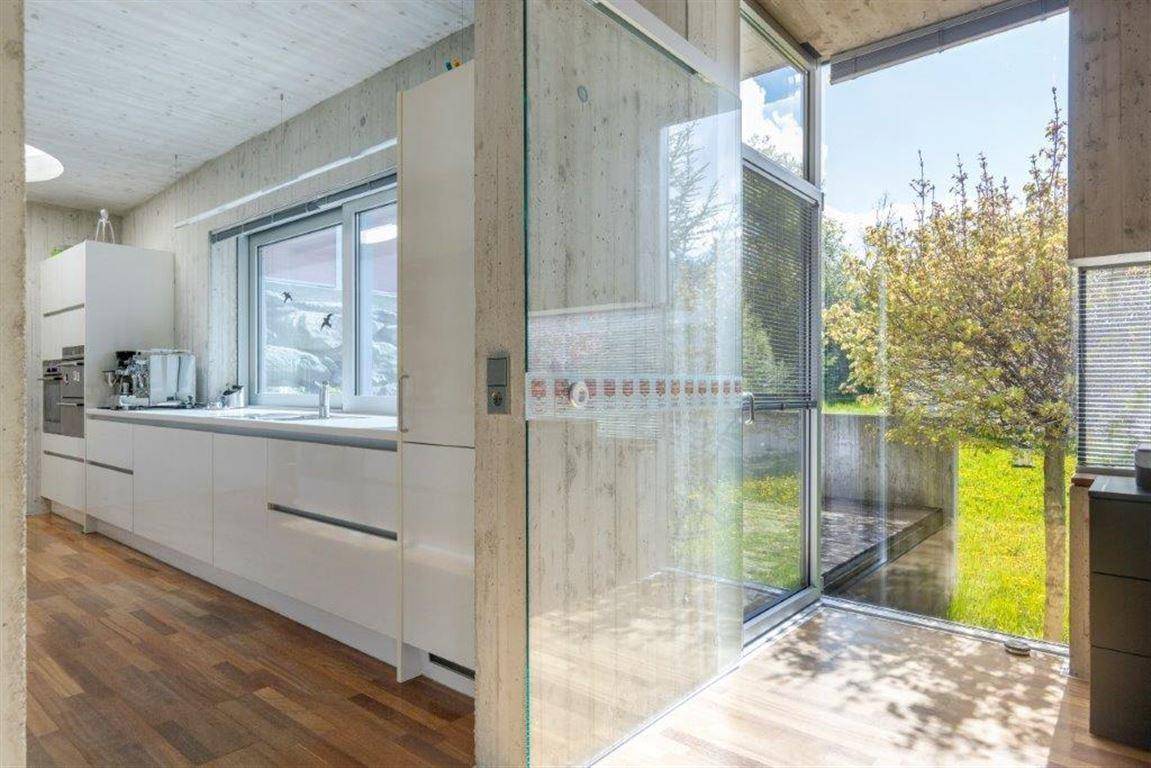 Moderne Küche mit weißen Schränken, Holzboden und großen Fenstern mit Blick ins Grüne.
