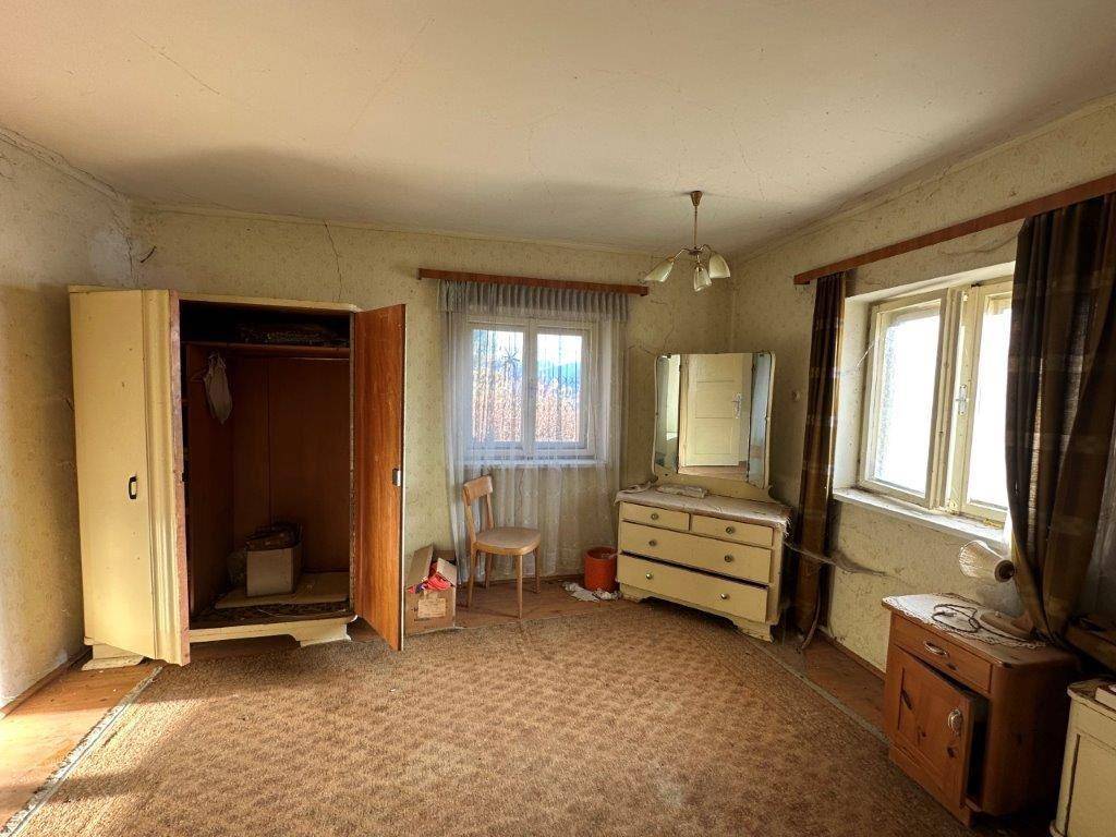 Schlafzimmer mit Teppichboden, Kommode und Fenster, das viel Licht hereinlässt, renovierungsbedürftig.
