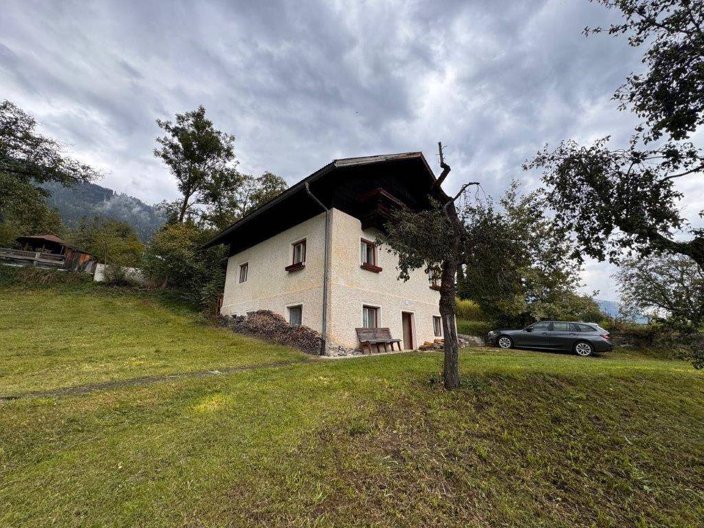 Ländliches Haus mit heller Fassade und Holzbalkon, umgeben von Wiesen unter bewölktem Himmel.