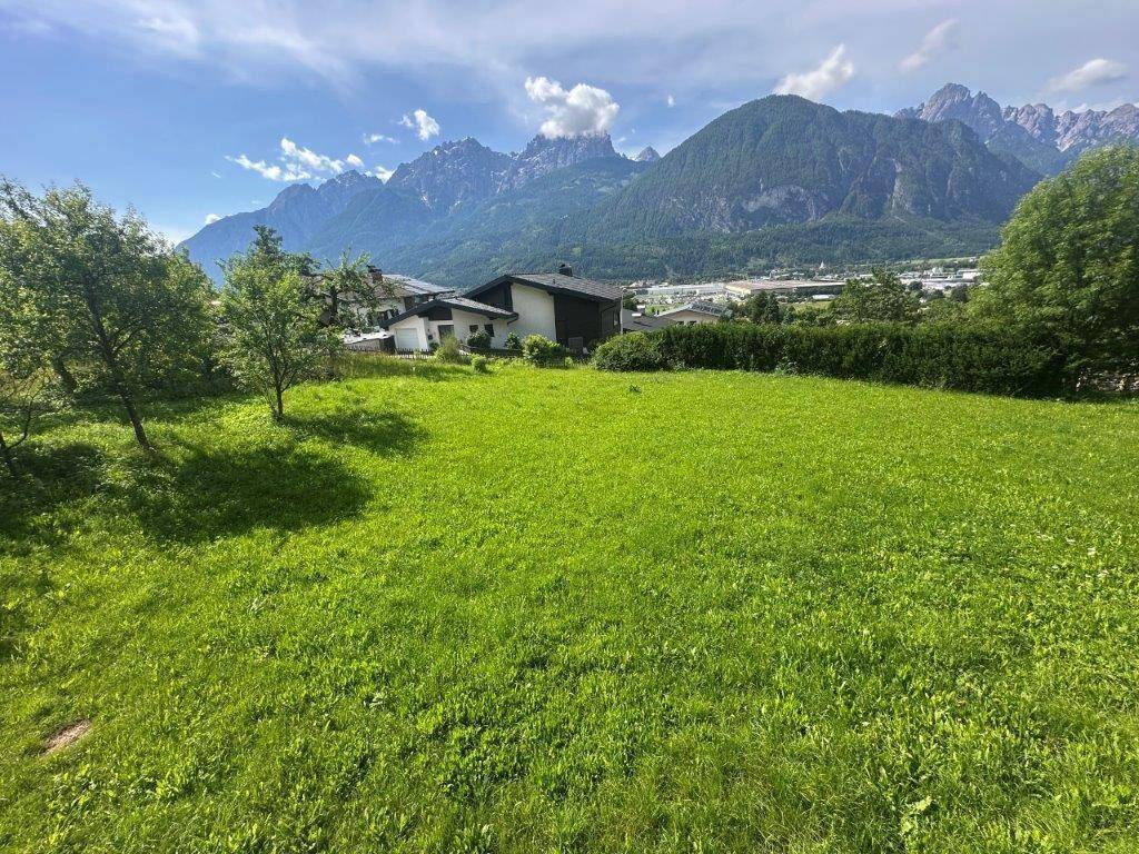 Idyllischer Ausblick über eine grüne Wiese auf eine Siedlung und die umliegenden Berge.