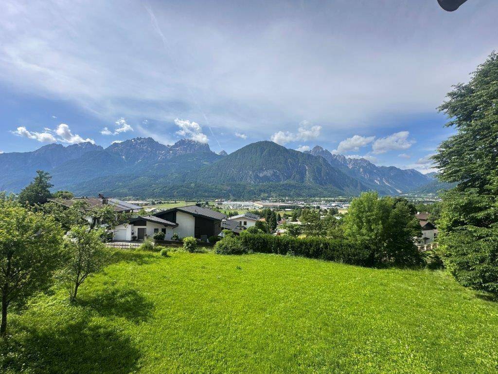 Weitläufiger Blick über grüne Wiesen und eine Siedlung mit majestätischen Bergen im Hintergrund.