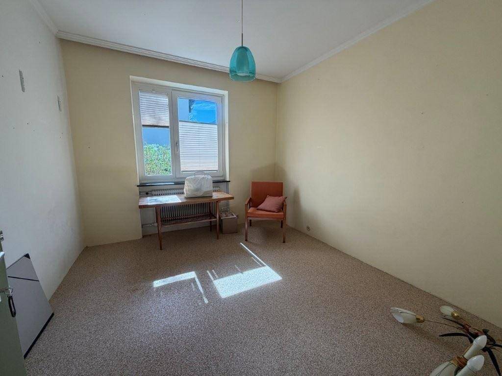Helles Zimmer mit Teppichboden, großem Fenster und einfacher Einrichtung.