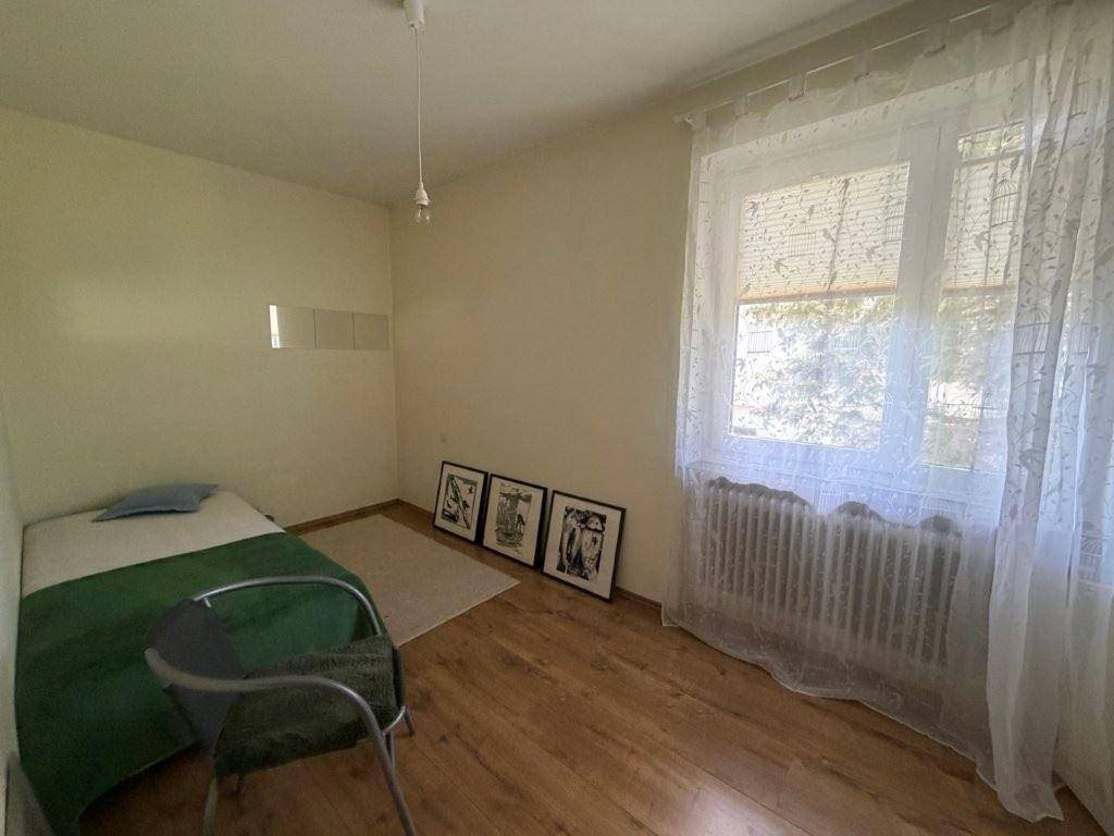 Einfaches Schlafzimmer mit Holzboden, Einzelbett und Fenster mit Vorhängen.