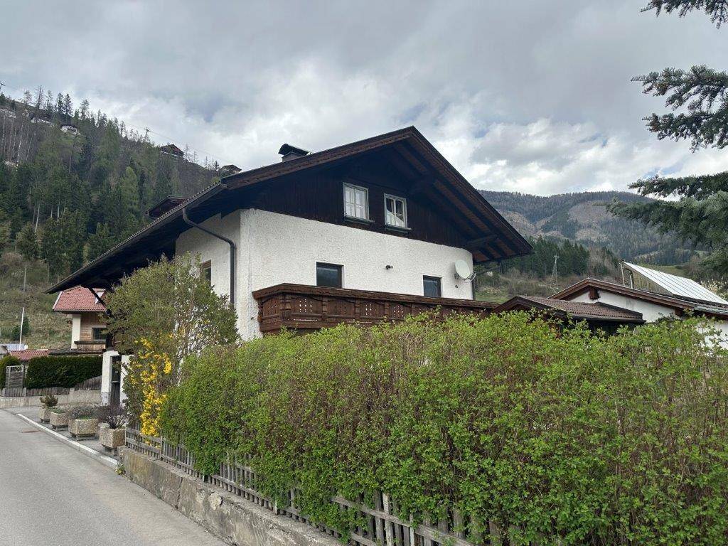 Ansicht des Hauses von der Straße mit Balkon, grüner Hecke und Bergen im Hintergrund.