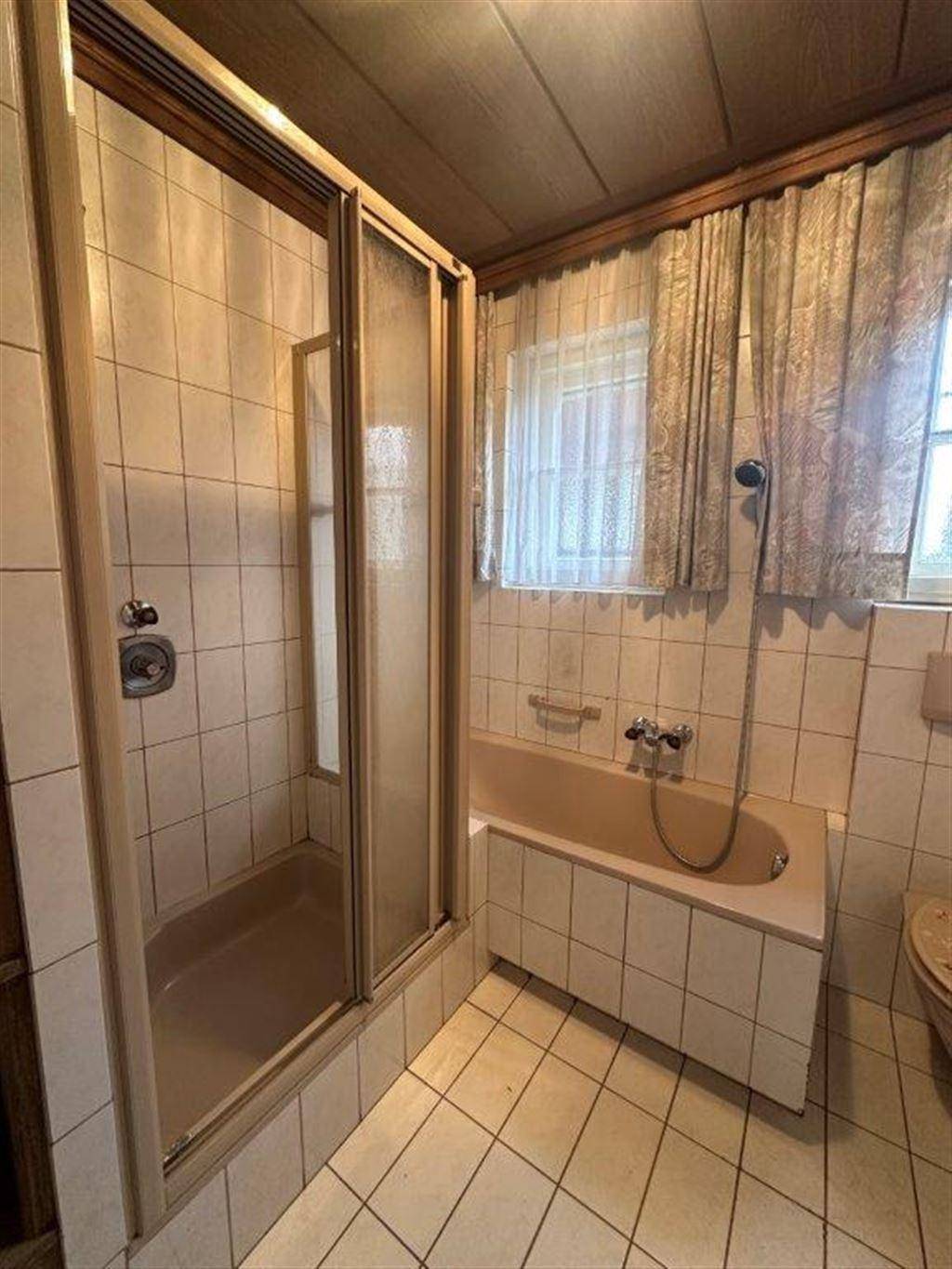 Badezimmer mit Duschkabine, Badewanne, beigen Fliesen und einem Fenster für Tageslicht.