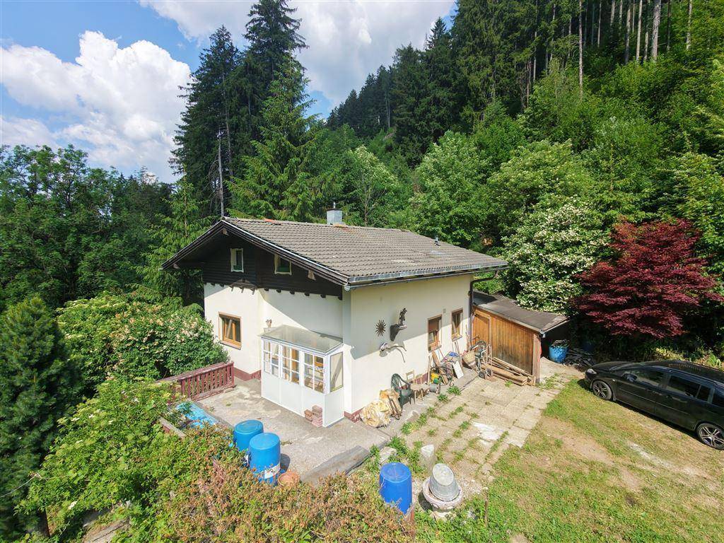 Haus in grüner Hanglage mit angrenzendem Schuppen und Parkplatz, umgeben von Wald.