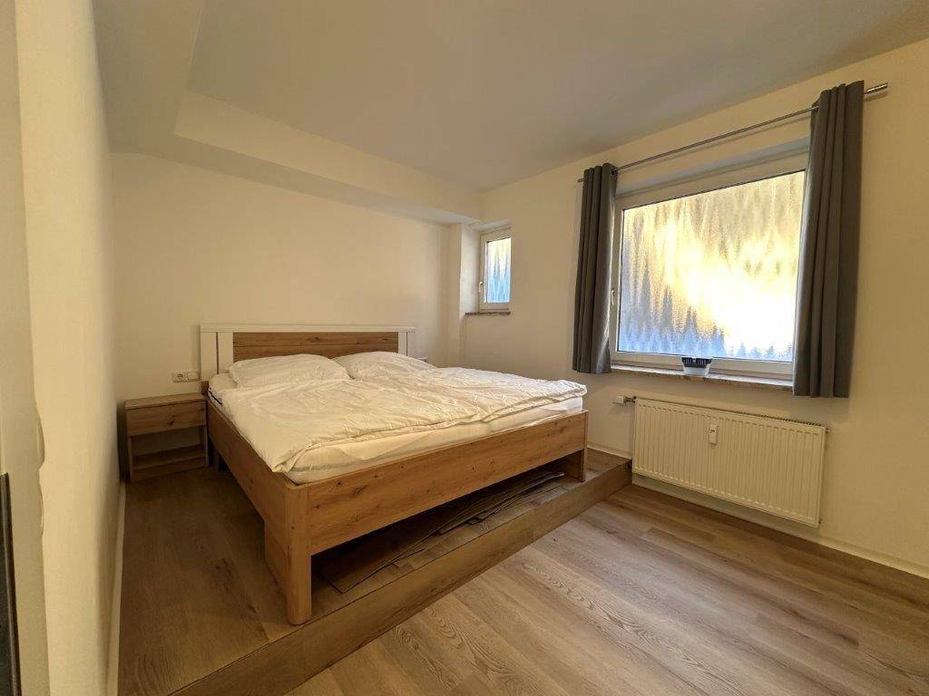 Schlafzimmer mit hellem Holzbett, Laminatboden und zwei Fenstern für natürliche Belichtung.