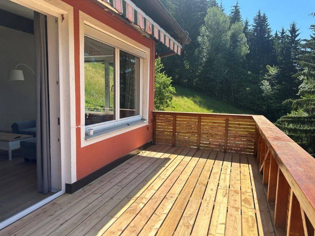 Heller Holzbalkon am roten Gebäude, umgeben von grünen Bäumen und Natur.
