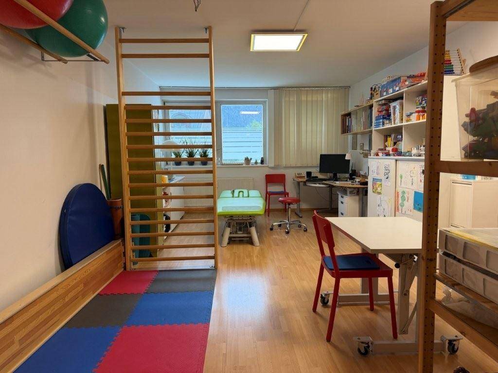 Therapieraum mit Holzfußboden, Sprossenwand, Behandlungsliege und Blick ins Grüne.