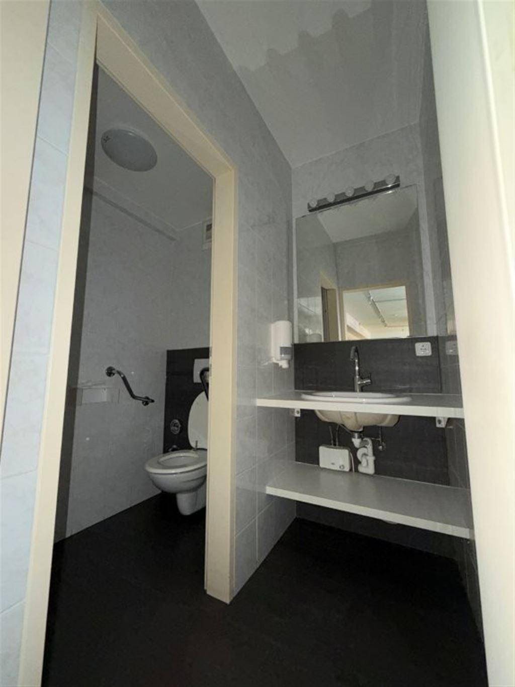 Badezimmer mit Fliesenboden, Waschbecken, Spiegel und Haltegriffen an der Toilette.