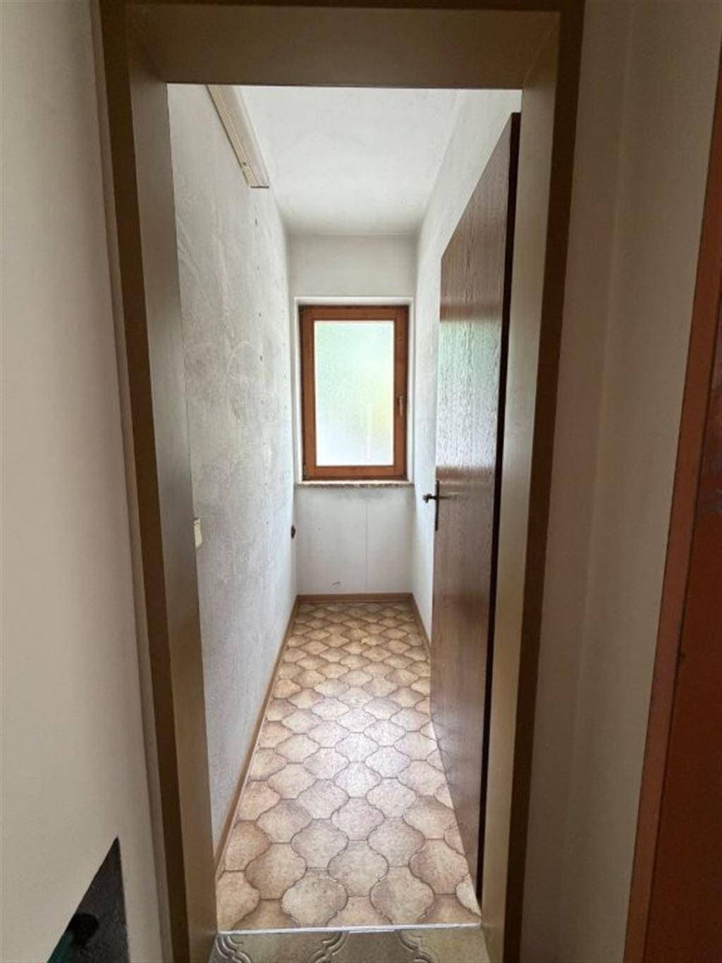 Schmaler Flur mit Fliesenboden und Fenster am Ende, der zu weiteren Räumen führt.