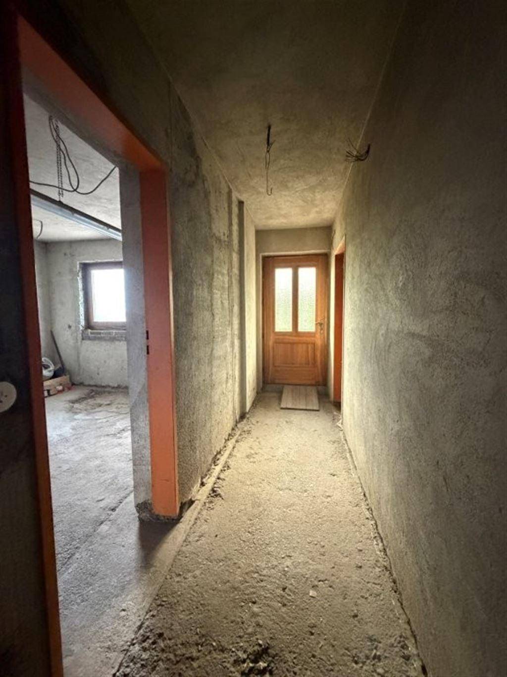 Flur im Rohbau mit Betonwänden und einer Holztür, die zu einem weiteren Raum führt.