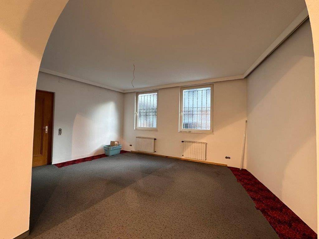 Geräumiger Innenraum mit Teppichboden, zwei Fenstern mit Gitterstäben und einem Durchgang zu einem weiteren Bereich.