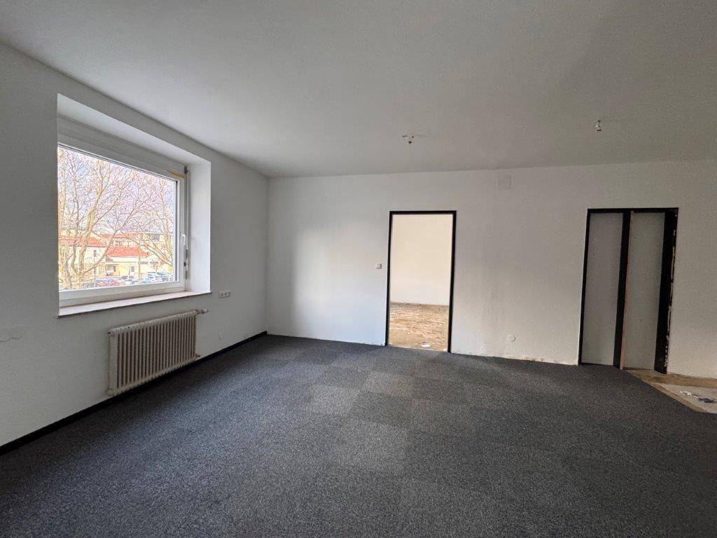 Großer Büroraum mit Teppichboden und einem Fenster, das Tageslicht hereinlässt.