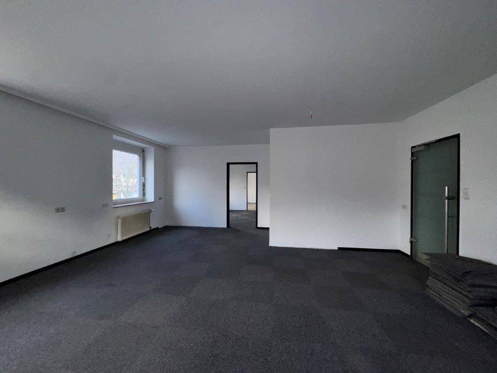 Heller Büroraum mit Teppichfliesen und Fenster, der Zugang zu weiteren Räumen bietet.