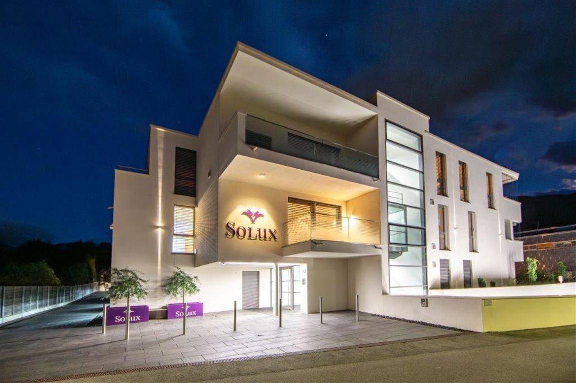 Moderne Wohnanlage bei Nacht mit beleuchteter Fassade und "Solux"-Logo am Eingangsbereich.