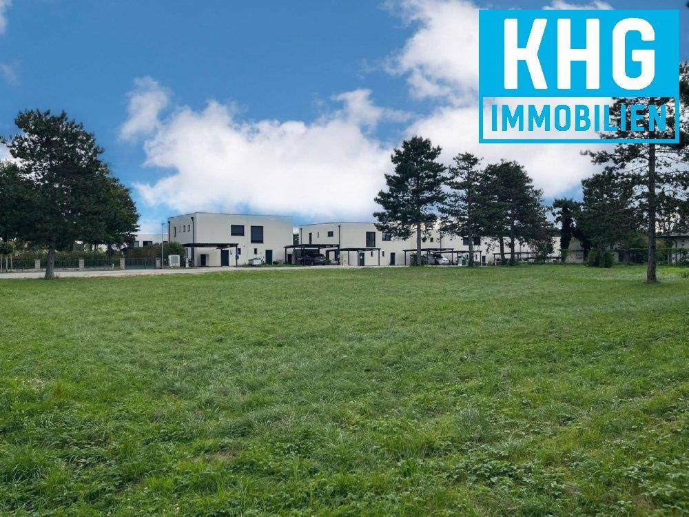 Offene Grünfläche mit modernen Gebäuden und Bäumen im Hintergrund unter blauem Himmel.