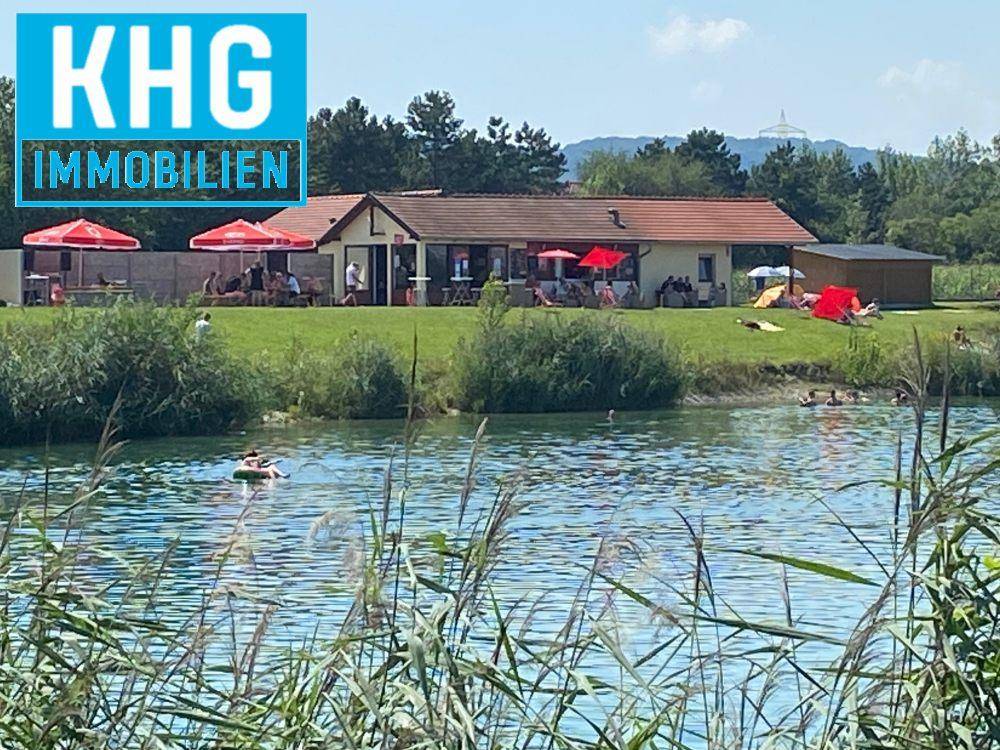 Blick auf einen Badesee mit schwimmenden Personen und einem Gebäude mit Sonnenschirmen am Ufer.