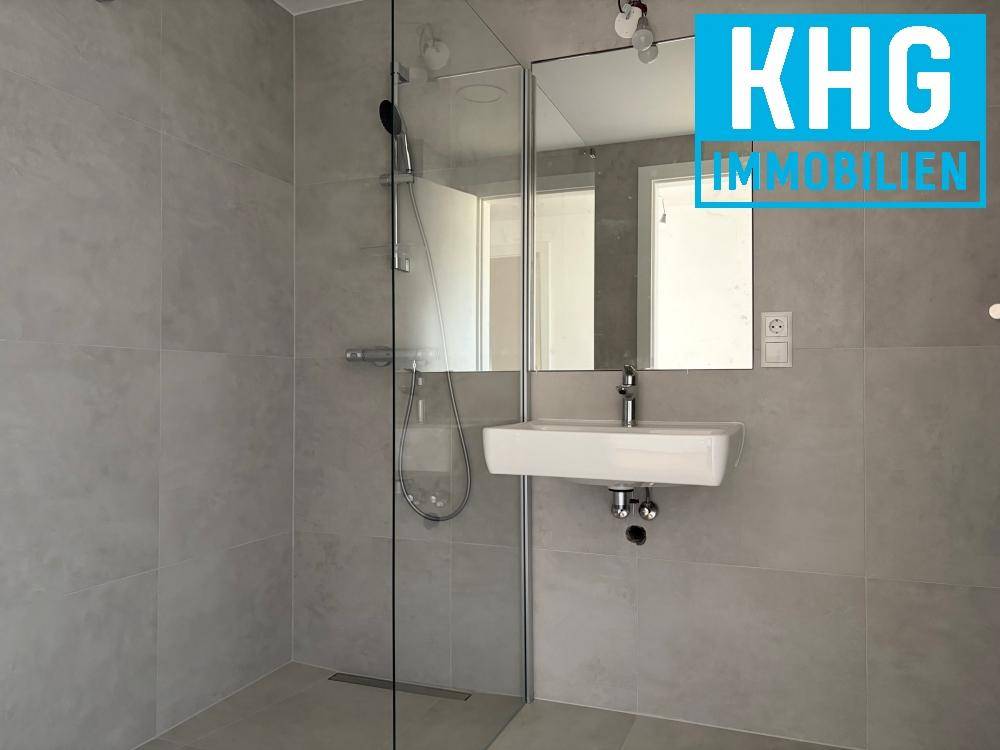 Modernes Badezimmer mit ebenerdiger Dusche, kleinem Waschbecken und grauen Fliesen.