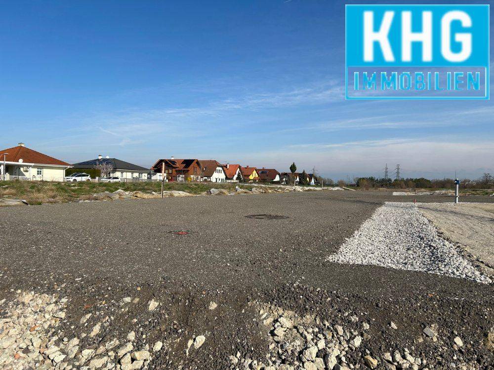 Bauland mit Schotterfläche und neuen Häusern im Hintergrund unter klarem Himmel.